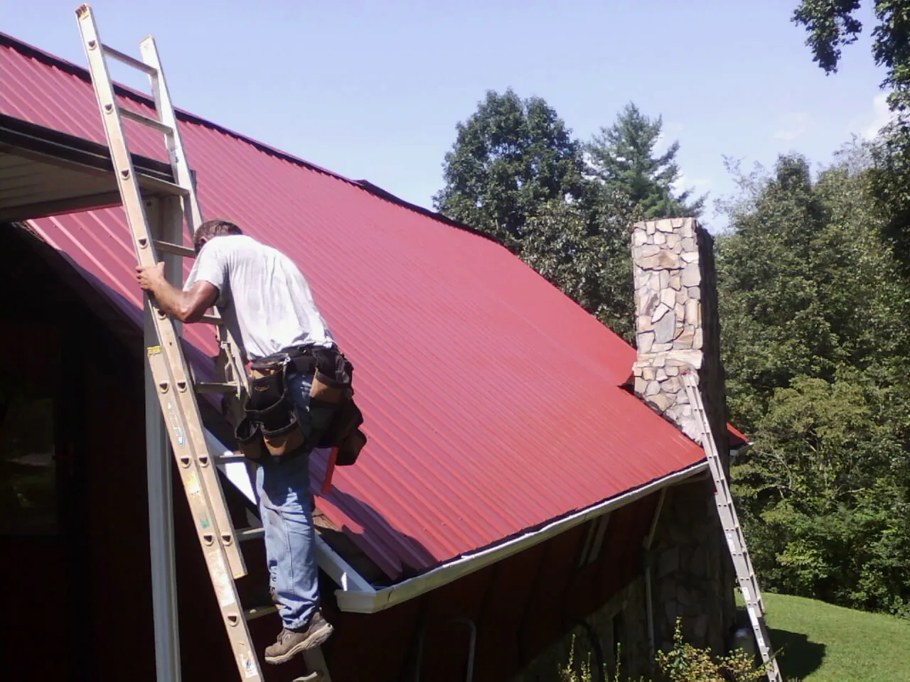 Roofer on a ladder beside a red metal roof and stone chimney — Soffit & Fascia Repair by Roofing Brothers in Umpqua